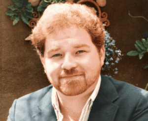 A smiling man with curly red hair and a beard, wearing a suit jacket.